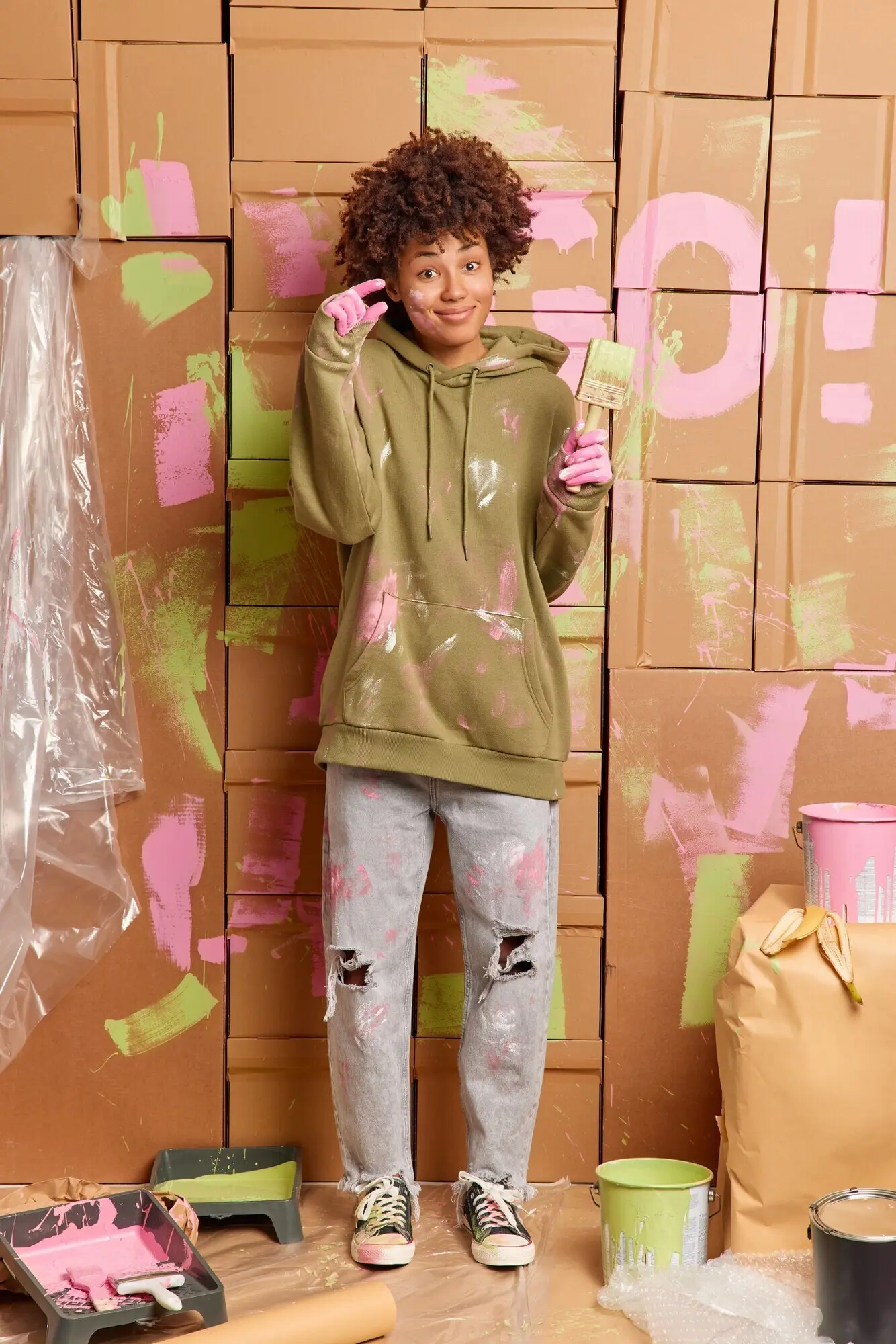 Ethnische Malerin macht eine kleine Geste und sagt, sie brauche nicht viel Zeit, um die Renovierung der Wände abzuschließen; hält einen Pinsel, steht in einem unordentlichen Raum, dekoriert die Wohnung und arbeitet an einem modernen Design. Neugestaltung.